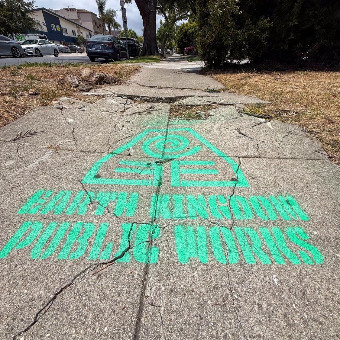 Een groene stencil met Earth Kingdom Public Works en het embleem van het Earth Kingdom, gespoten op een gebarsten stoep in Los Angeles