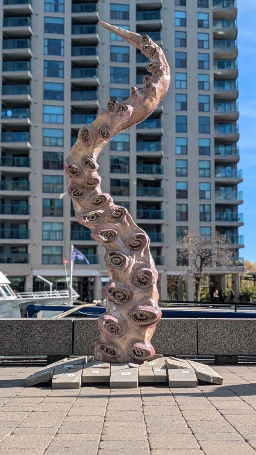 The printed tentacle, 3 metres tall, rising out of broken paving slabs on Toronto's waterfront, a residential tower and boats behind