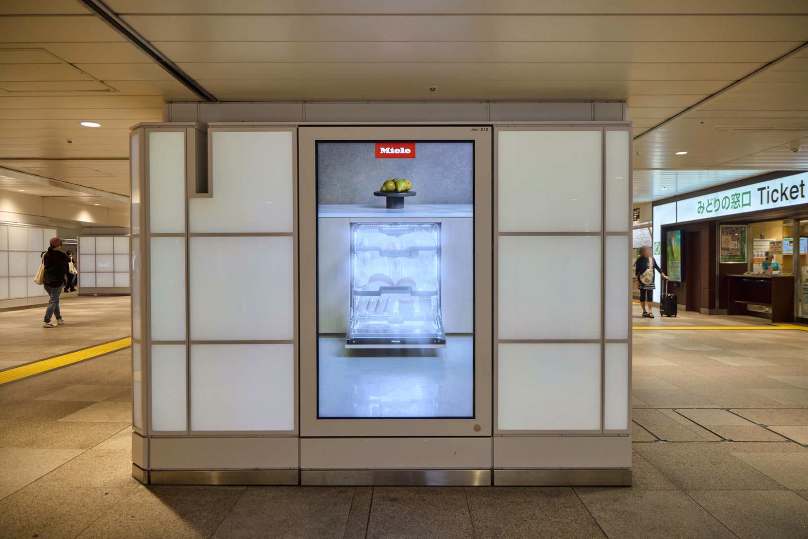 A J-AD Vision pillar screen showing the Miele dishwasher glowing white, next to the JR ticket office