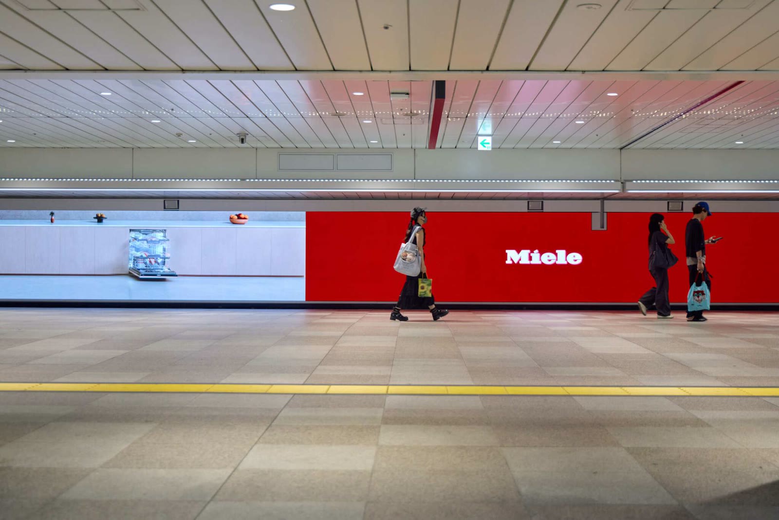 Shinjuku Wall 456: the white Miele kitchen with a dishwasher open, next to the red Miele logo block, passers-by in front